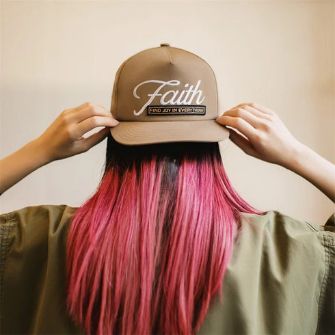 Person wearing a cap with 'Faith' text and pink hair against a beige background